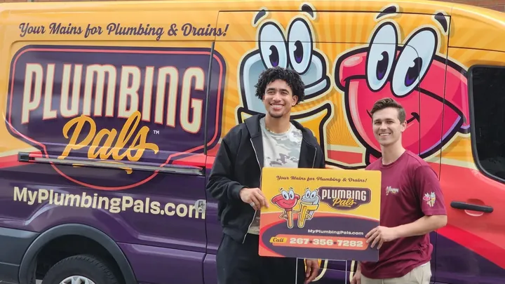 Two Plumbing Pals technicians holding the company sign in front of a wrapped van